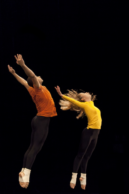 Two dancers in mid air. They arch their upper backs with their hands extending to the ceiling